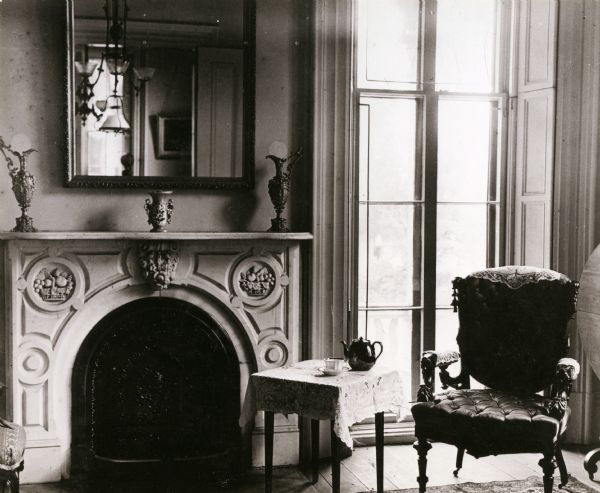 Interior of the parlor. The fireplace is on the left, with a mirror above the mantel. A large window at floor height is on the right behind a chair and a small table.