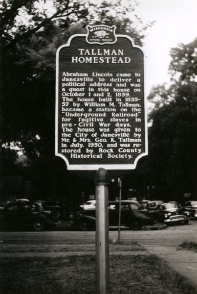 Tallman Homestead Marker. There are automobiles parked along the curb in the background.