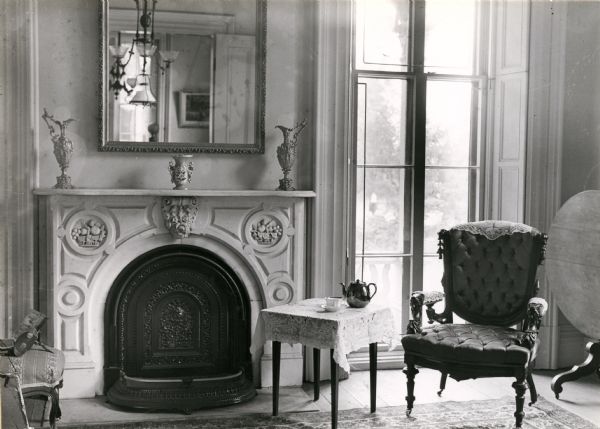 View of the second parlor in the Tallman House. The fireplace is on the left, with a mirror above the mantel. A large window at floor height is on the right behind a chair and a small table. Another tilt top table is on the far right, tilted up.