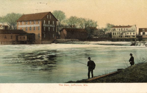 View towards a dam in Jefferson. Two men are fishing from the shoreline in the foreground. The Woolen Mills are on the opposite shoreline. Caption reads: "The Dam, Jefferson, Wis."