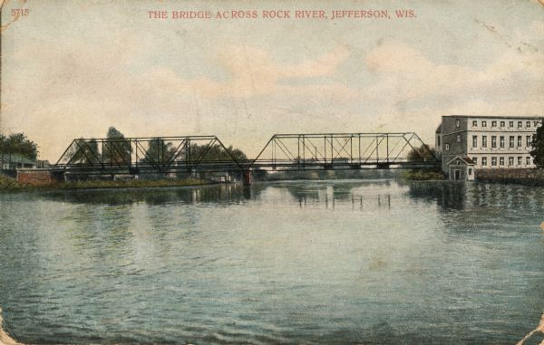 View across water towards the bridge across the Rock River. An industrial building is to the right of the bridge. Caption reads: "The Bridge Across Rock River, Jefferson, Wis."