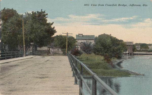 View down Crawfish Bridge, with a horse-drawn buggy approaching the bridge. Buildings and trees are along the river on the right. Caption reads: "View from Crawfish Bridge, Jefferson, Wis."