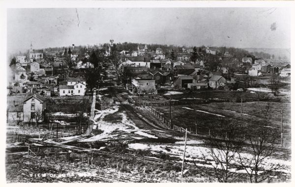Aerial view with the main street with railroad tracks in the foreground and houses in the background. There is also a church in the upper left corner.