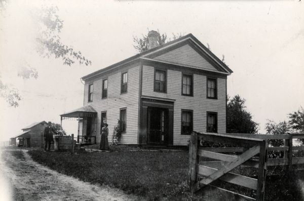 View up road towards the residence of L.S. Fowler. A man on the left is petting a dog that is sitting on some type of platform, and a woman is standing in the center near the corner of the house. In the background on the left an automobile is parked next to a building under a roof.