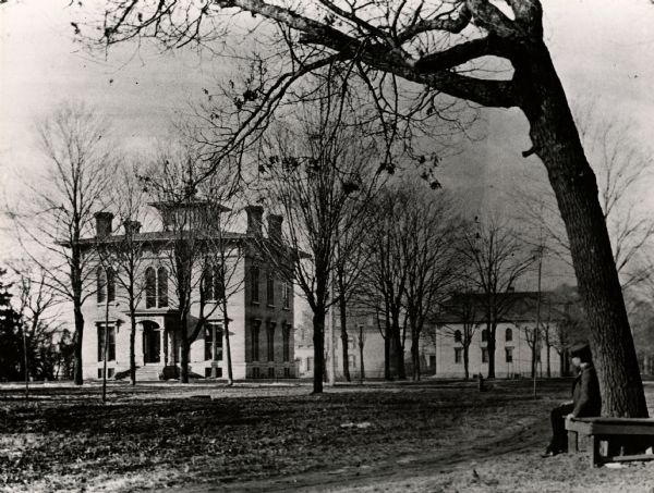 The residence of O.S. Head. A man is sitting in the foreground on a wrap-around bench around a tree.