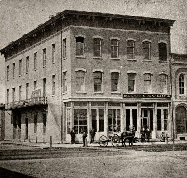 View across intersection towards Kimball's Hall, one of the first brick buildings in Kenosha. A sign on the right side of the building reads: "Dewey & Sinclair", and two men are standing under the sign in front of the entrance. Three other people are standing on the sidewalk at the corner of the building near a horse-drawn vehicle.