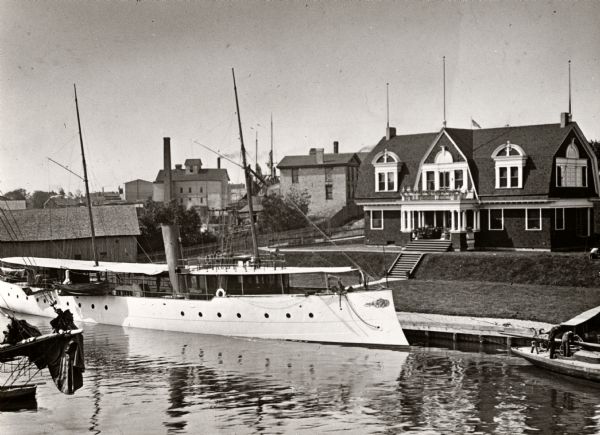 The Morgan Club House on the river. A long, narrow excursion boat is moored on the dock in front of the club.