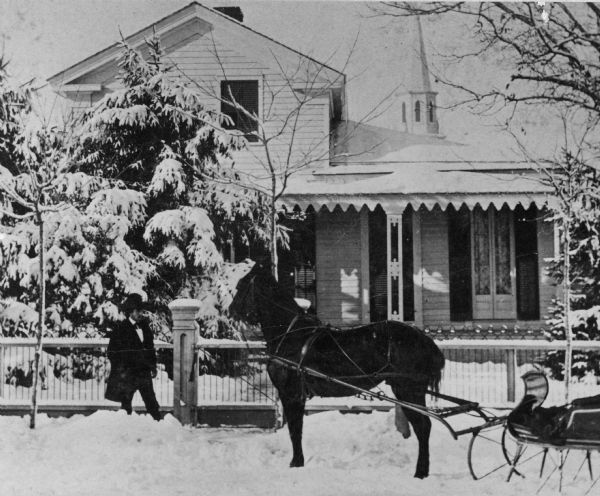 The home of photographer S.W. Truesdell. A man is standing in the snow in front of the fence near a horse and sleigh.