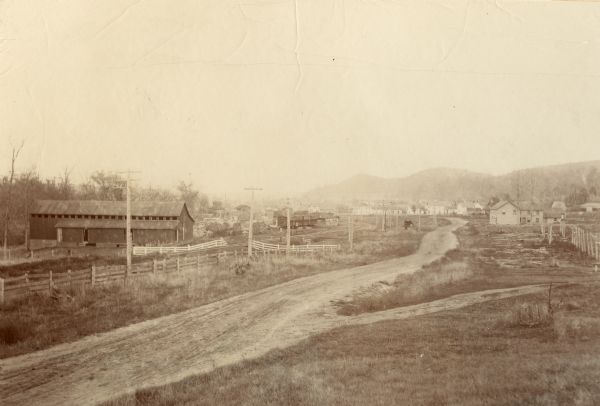 Road lined with fences leading into town. Several houses are visible in the distance. A railroad track and electric or telegraph line runs along the left side of the road.