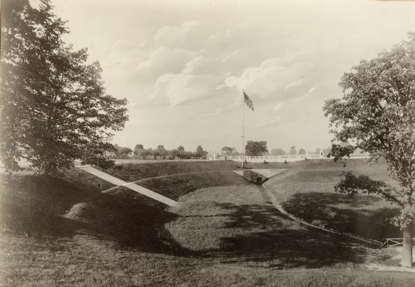 View of Kohler Ravine Park. In the right foreground is a small bridge, and on the right is a set of wide, concrete steps leading from the road down the hill to the narrow channel of water at the bottom. In the background is a flagpole with a flag in front of a bridge with columns at each end and a guard rail between.