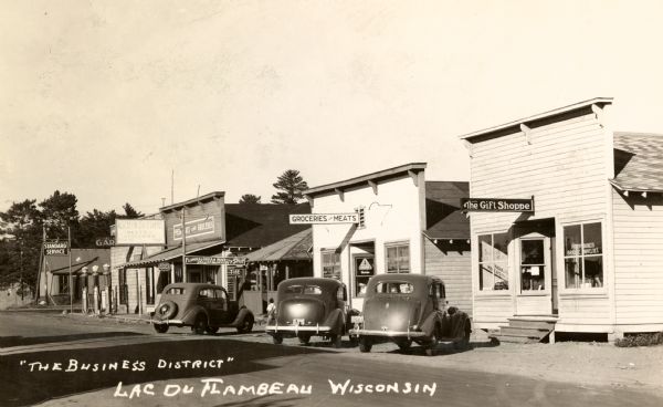 View from street towards cars parked at an angle in front of storefronts. Signs on the buildings read: "The Gift Shoppe," "Groceries and Meats," "Flambeau Indian Novelty Shop Genuine Handicraft," Aschenbreners Meats & Groceries" and "Filling Garage." Caption reads: "'The Business District' Lac du Flambeau, Wisconsin".