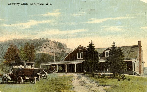 View up dirt road toward club. Automobiles are parked on the left. There is a bluff in the background. Caption reads: "Country Club, La Crosse, Wis."