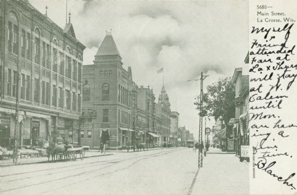 View down right side of street towards the left. Pedestrians and horse-drawn vehicles are along the sidewalks and curb. Caption reads: "Main Street, La Crosse, Wis."