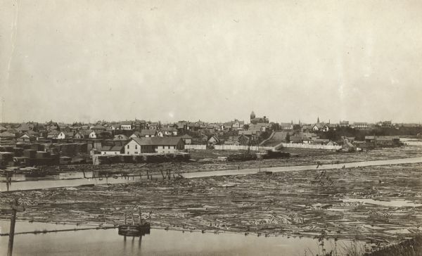 Elevated view across river towards the town. Caption reads: "Ladysmith, Wis." Logs are floating in the water in the foreground.