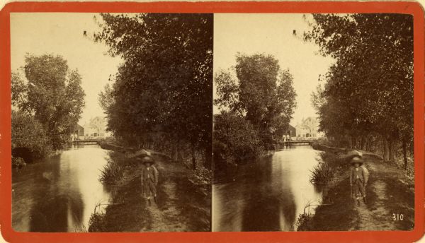 Stereograph of a little boy with a straw hat and stick walking along an outlet into Lake Geneva.