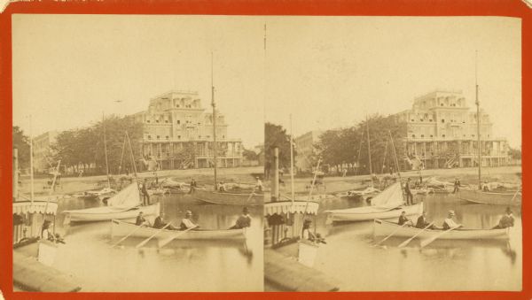 Stereograph view of the Whiting House with its large harbor and boats. Whiting House is a four-story wood frame Second Empire style hotel standing on the lake shore at Lake Geneva. Several sets of stairs lead to a porch which extends across the front of the first floor. Several rowboats and sailboats are moored in front of the hotel. Horse-drawn vehicles are parked on the shore. Many people can be seen standing on land or aboard the boats. In the foreground, four people are seated in a rowboat, three are rowing, and one is seated in the stern. Other buildings of the village are seen on the left.