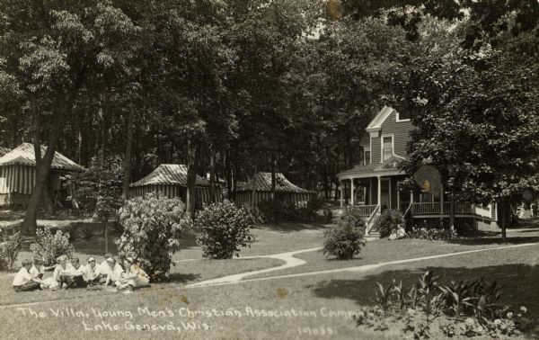 The villa at the Young Men's Christian Association Camp. A group of girls and boys are sitting on the lawn on the left and two more people are sitting on the lawn near the building on the right. Striped tents are in the background under trees. Caption reads: "The Villa, Young Men's Christian Association Camp, Lake Geneva, Wis."