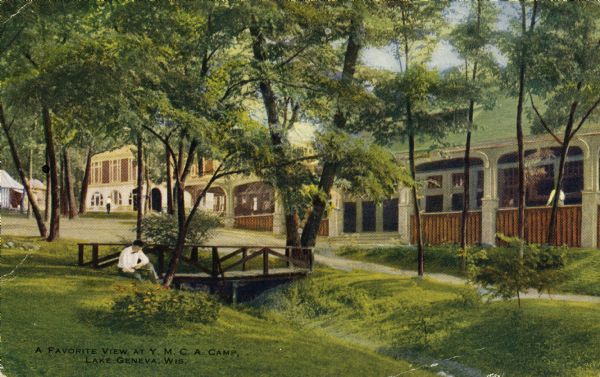 The Young Men's Christian Association Camp. A man is sitting on the lawn near a small bridge on the left. In the background along the right are buildings. Caption reads: "A Favorite View at Y.M.C.A. Camp, Lake Geneva, Wis."