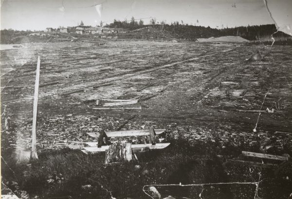 Lake Shore Lumber Company of Oshkosh's mill waste lumber slab dump. Leander Choate, owner of mill, used this marsh for his waste lumber which was piled at least 8 feet high (most of it below water level). Most mills used this waste for fuel, and why Choate didn't is an undetermined fact; perhaps this was surplus above the fuel needs. Note the track across the marsh for transport. Piled remnants are still there (1960) and are rotting as false "ground" fill. Cottages are now built on top of this questionable foundation. In the background is the town of Lake Tomahawk which was just beginning to look to tourist trade as the lumber was depleted.