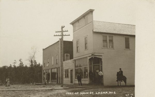 View across Main Street in Laona. Three men are standing in front of the corner building, and another man is on horseback to the right. On the left in the background is a woman carrying a child. Caption reads: "Part of Main St. Laona, Wis."