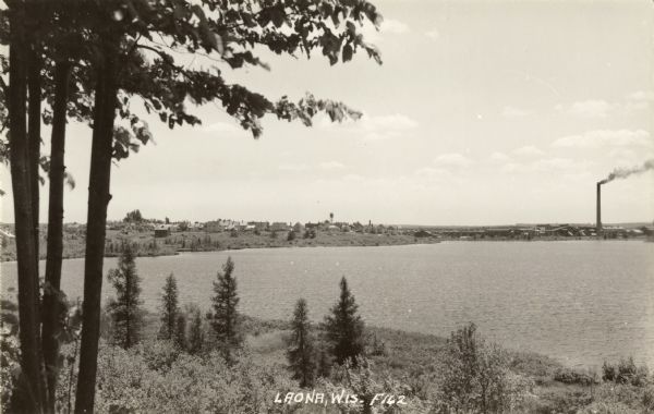 Elevated view from hill towards the city of Laona on the far shoreline. A large smokestack is spewing smoke. Caption reads: "Laona, Wis."