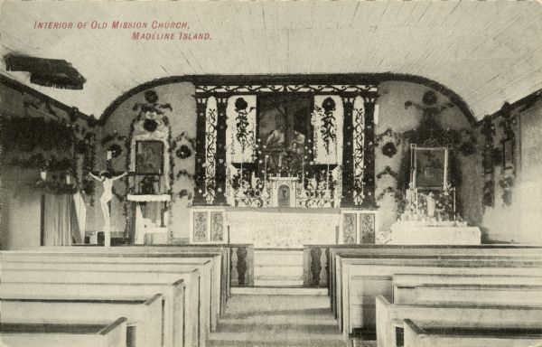 The interior of St. Joseph's Catholic Church. It was built in 1841 by Father Frederick Baraga (later Bishop), who established his mission in 1835. A small log church was commenced the same year. Later when a larger church was needed, this one was re-erected on a different site. Contrary to local legend, this is not on the site of the mission established by Fr. Claude Allouez in 1665. His mission, La Pointe du Sainte Esprit, was built on the mainland near or in the present ciy of Bayfield, Wisconsin. The altar appointments are Roman Catholic in iconography. Caption reads: "Interior of Old Mission Church, Madeline Island."