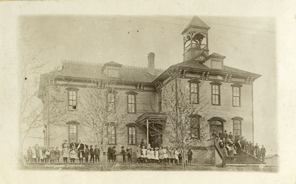 A high school in Linden. Students and teachers are standing along the sidewalk, and another group of people are on the front steps in front of the building.