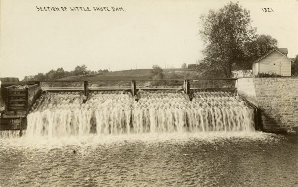 View across water looking towards the dam. There is a stone wall to the right of the dam, and a building above it. Caption reads: "Section of Little Chute Dam." A hill is in the background.