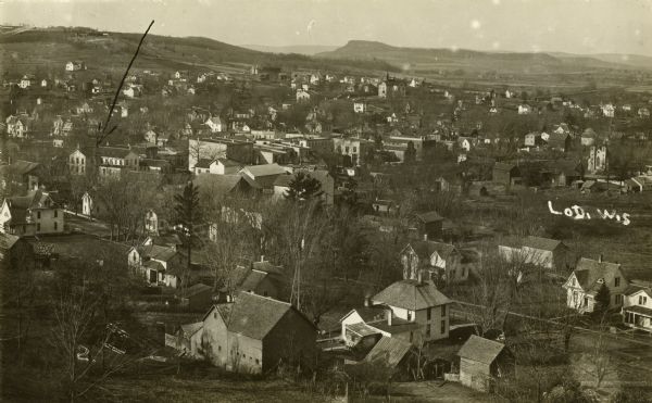 Elevated view of a residential neighborhood, with some larger buildings. An arrow has been drawn on the print pointing to a building on the left. Hills are in the distance.