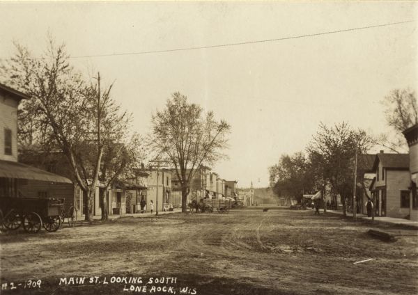 Text on front reads: "Main Street Looking South, Lone Rock, Wis." Unpaved street with sidewalks, dwellings and buildings on both sides. Horse drawn vehicles are hitched at the curb and pedestrians are on the sidewalk.