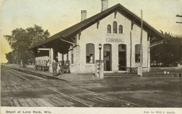 View across street and railroad tracks toward the railroad depot in Lone Rock. A man is standing on the platform. Caption reads: "Depot at Lone Rock, Wis."
