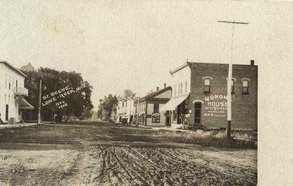View down unpaved street in Lone Rock. A building on the right has a sign painted on the side that reads: "Monona House". Caption reads: "St. Scene Lone Rock, Wis."