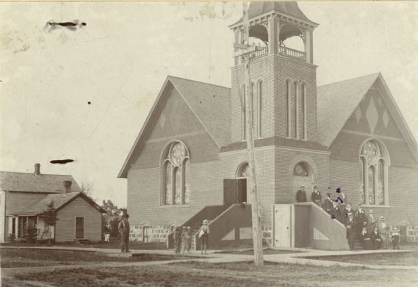 Methodist Episcopal Church and parsonage built under Rev. G.N. Foster. Groups of people are standing on the entrance steps, and others are standing on the sidewalk.