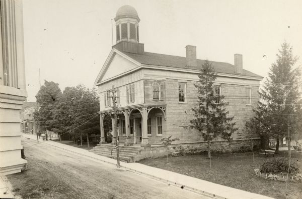 Slightly elevated view across street towards the second Iowa County Court House, built in 1842. For a time in 1846, the space was leased to the newly organized congregation of Trinity Episcopal Church while its own edifice was under construction. After the seat of county government was moved to Dodgeville in 1861, the city of Mineral Point purchased the property. The building was demolished in 1913 and the Municipal Building was erected on its site.