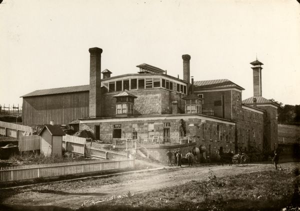 Mineral Spring Brewery, located on 272 Shake Rag Street, erected in 1850 (currently 276 Shake Rag Street). A group of four men are posing outdoors on the road at the corner of the building, and another person is sitting in a horse-drawn carriage on the right.