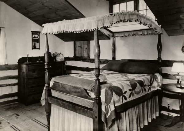 A bedroom in the restored Cornish miner's house, called Polperro, on Shake Rag Street. The house was temporarily covered with siding, but after restoration the siding was removed to reveal the original log construction. The building is currently owned by the Wisconsin Historical Society and is one of several structures that comprise the Pendarvis historic site. The room features a four-poster bed covered with a quilt and a high dormer window set into the sloped ceiling.
