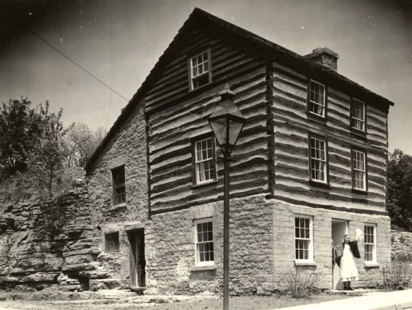 A woman demonstrating how Shake Rag Street got its name, with the restored Cornish miner's house, called Polperro, behind her. The house was temporarily covered with siding, but after restoration the siding was removed to reveal the original log construction. The building is currently owned by the Wisconsin Historical Society and is one of several structures that comprise the Pendarvis historic site.
