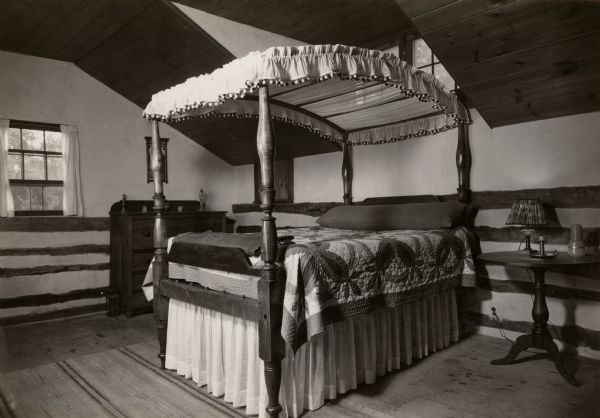 The bedroom in the restored Cornish miner's house, called Polperro, on Shake Rag Street. The house was temporarily covered with siding, but after restoration the siding was removed to reveal the original log construction. The building is currently owned by the Wisconsin Historical Society and is one of several structures that comprise the Pendarvis historic site. The room features a four-poster bed covered with a quilt and a high dormer window set into the sloped ceiling.
