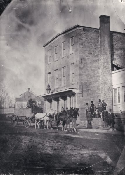 The U.S. Hotel was constructed in 1852 by Hugh Phillips to be used for mercantile purposes, but later transformed into the hotel. A group of people are standing near the hotel, and a team of horses are pulling a wagon on the street.