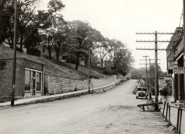 View up Main Street, with houses up on a hill on the left above a stone wall that runs along the sidewalk. On the right is a sign that reads: "Maiden R[ock] Post O[ffice]", and another just behind it that reads: "Ben's Tavern". 