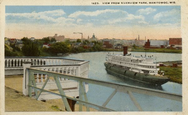 Elevated view of Manitowoc from Riverview Park. A large boat is in the bend of the river. In the foreground is a railing and an extended overlook over the river. Caption reads: "View from Riverview Park, Manitowoc, Wis."