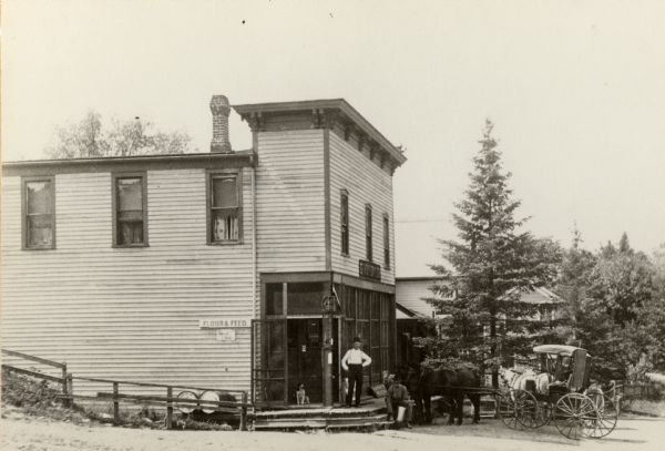 Two men and a horse and carriage outside of Thompson Store. A dog is sitting in the open doorway at the corner of the building. A sign on the side reads: "Four & Feed".