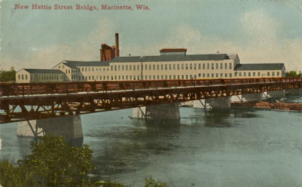 View from shoreline toward the new Hattie Street Bridge. Factory buildings are on the shoreline behind. Caption reads: "New Hattie Street Bridge, Marinette, Wis."