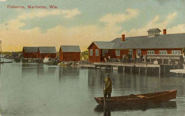 View across water toward buildings on the right, with men standing on a platform at the shoreline in front of a large building. Three more smaller buildings are further down the shoreline. A man is standing in a rowboat in the foreground. Caption reads: "Fisheries, Marinette, Wis."