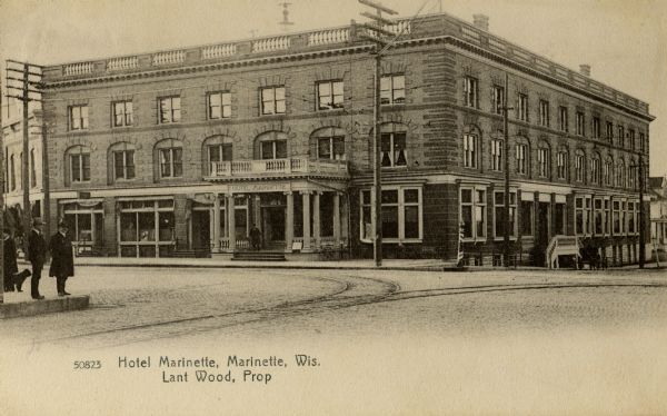 View across intersection toward the hotel. Standing on the sidewalk at the corner on the left are men and a dog. Caption reads: "Hotel Marinette, Marinette, Wis. Lant Wood, Prop."