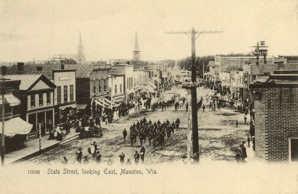 Elevated view of State Street. There is a parade coming down the street, and crowds are on the sidewalks. Caption reads: "State Street, Looking East, Mauston, Wis."