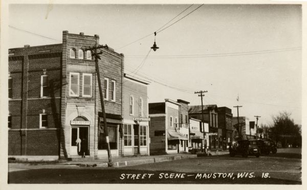 View across intersection towards storefronts on the left. On the corner is the Post Office, and further down are signs for the "Exide Battery Station," and "Buck Inn -- Eat." Automobiles are parked at an angle along the curb. Caption reads: "Street Scene -- Mauston, Wis."