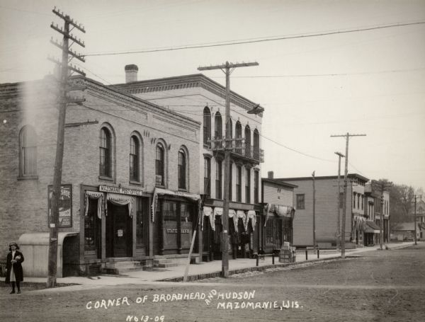 Mazomanie Post Office and the State Bank on the corner of Broadhead and Hudson. On the left a young girl stands in the dirt road. Further down the block a young child is on the sidewalk in front of a store displaying coats and jackets.