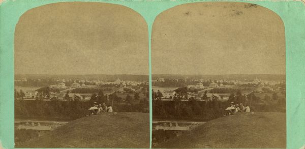 Stereograph view of Menomonie. A group of people are sitting on a hill in the foreground overlooking the town.