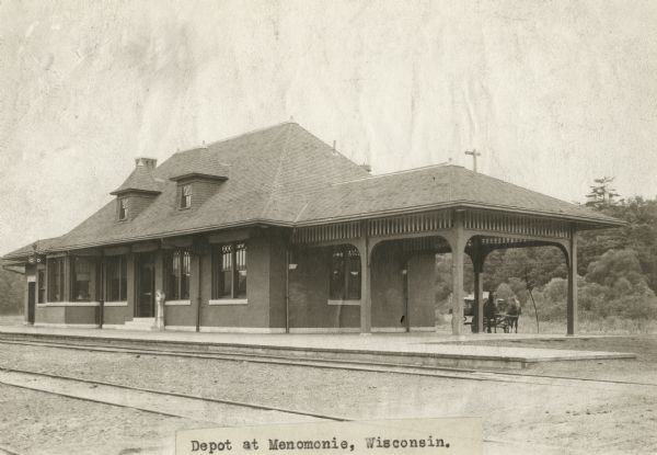 Exterior of railway depot. A horse-drawn vehicle is parked behind the station on the right.